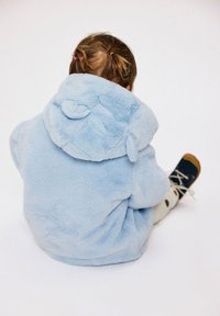 Šviesiai mėlyna, pūkuota džemperio striukė su gobtuvu ir apvaliais ausytėmis ant gobtuvo. Vaikas sėdi, dėvi tamsius batus su raišteliais ir minkštas smėlio spalvos kelnes.