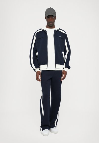 Navy tracksuit with white side stripes, featuring a zip-up jacket and matching pants, complemented by a white t-shirt and gray baseball cap.