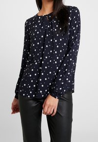 Blusa blu navy con motivo a stelle bianche, con fronte plissettata, scollo rotondo e maniche lunghe arricciate, abbinata a pantaloni neri simili alla pelle.
