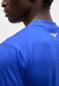 Κοντινό πλάνο ενός ατόμου που φοράει μπλε πουκάμισο με μικρό λευκό λογότυπο Mizuno κοντά στη λαιμόκοψη στην πλάτη.