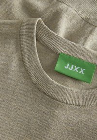 Beżowy sweter z dzianiny z okrągłym dekoltem i ściągaczami, z zieloną metką z napisem "JJXX". Miękka tekstura i gładkie wykończenie.