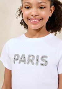 Biała bawełniana koszulka z teksturowanym srebrnym napisem "PARIS" ozdobionym przezroczystymi cyrkoniami, okrągłym dekoltem i krótkimi rękawami.