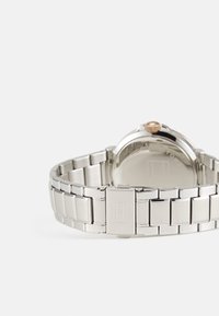 Montre en acier inoxydable argenté avec un bracelet en métal lisse. Le boîtier rond est doté d'une couronne en or rose ; logo minimal gravé sur le fermoir.