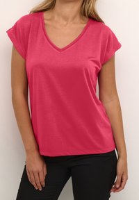Mujer vestida con una blusa de cuello en V rosa brillante y pantalones negros, de pie contra una pared clara y simple.