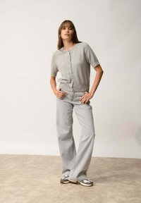 Szary kardigan z krótkim rękawem i guzikami, nałożony na szare szerokie dżinsy. Modelka w wzorzystych trampkach, pozująca na neutralnym tle.