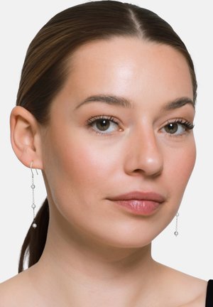 Kobieta o jasnej cerze i brązowych włosach zaczesanych do tyłu, nosząca delikatne srebrne kolczyki z małymi białymi perłami na jednolitym białym tle.