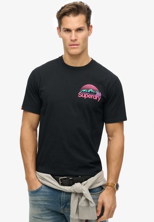 Sort bomulds T-shirt med korte ærmer, der har et grafik af bjerg i pink og blå samt "Superdry" tekst, parret med jeans og en grå sweatshirt.