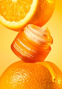 Orange jar of Ole Henriksen Banana Bright Eye Crème, featuring a smooth cream texture and translucent lid, surrounded by whole oranges on a yellow background.