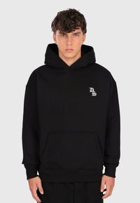 Jeune homme aux cheveux bouclés portant un sweat à capuche noir avec les initiales blanches "DS" sur la poitrine, se tenant devant un fond gris clair uni.