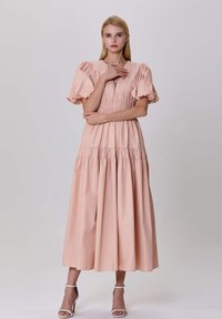 Robe rose pâle avec un corsage smocké, des manches bouffantes et une jupe à volants. Modèle portant des talons blancs à lanières, les bras croisés au niveau de la taille.