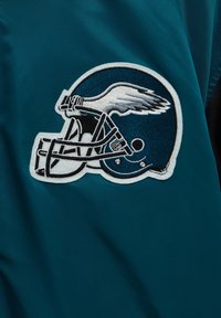 écusson brodé d'un casque de football bleu avec un détail d'aile blanche sur un fond en tissu sarcelle.