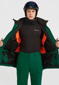 Skijakke i mørkegrøn med orange for, sort basislag og grønne skibukser. Har høj krave og foran lynlås design.