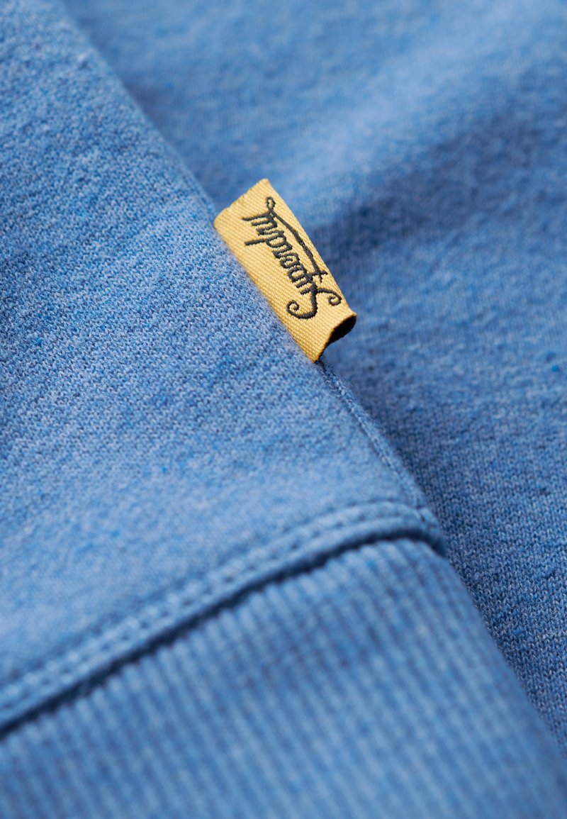 Sudadera azul hecha de tela suave, con puños acanalados y una etiqueta amarilla con texto negro cosida en el lateral.