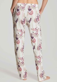 Vrouw in witte broek met roze en paarse bloempatronen, staande op blote voeten met handen in de zakken, van achteraf gezien.