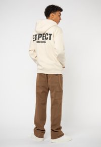 Cream hooded sweatshirt with "EXPECT NOTHING" printed on the back, paired with brown cargo pants. Material appears soft, casual style.