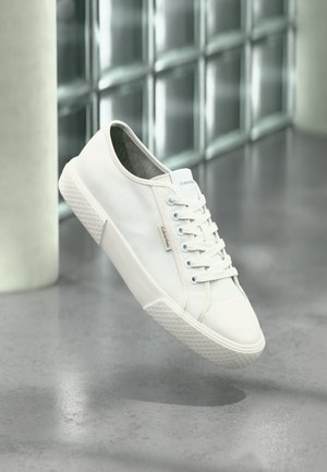 Biały niski sneaker Calvin Klein ze sznurowadłami, gumową podeszwą i metką z logo unoszący się nad szarą wypolerowaną podłogą w pobliżu ściany z bloków szklanych.