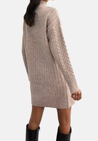 Žena v krémovém žebrovaném pleteném šaty se vzorem copánků na rukávech, v černých kotníkových botách, stojící s mírným otočením od objektivu.