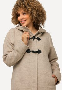 Cappotto in lana beige con una texture morbida, cappuccio e chiusure a moschettone. Presenta tasche laterali e una vestibilità rilassata, adatto per il tempo più fresco.