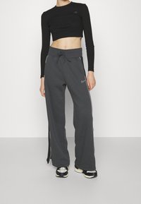 Osoba oblečená v černém dlouhém crop topu, tmavě šedých širokých teplácích Nike se bočními pruhy a černobílých teniskách, stojící proti bílé zdi.