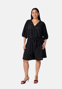 Vestido envolvente negro de manga corta, hecho de tejido texturizado, con detalles de ojales y un cinturón atado en la cintura, combinado con unos tacones rojos con tiras.