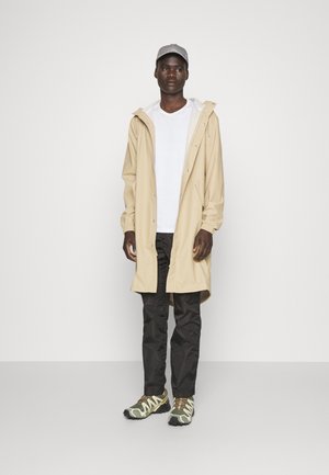 Beige regenjack met een capuchon, drukknoopsluitingen en elastische manchetten. Witte t-shirt eronder, zwarte broek en gestructureerde sportschoenen.