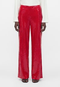 Personne portant un pantalon large à paillettes rouges brillantes, un haut blanc à manches longues et des sandales noires ouvertes, debout devant un fond uni.
