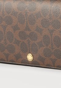 Καφέ υφασμάτινο πορτοφόλι από δέρμα με επαναλαμβανόμενο οβάλ μοτίβο και μικρό στρογγυλό χρυσό κουμπί με ανάγλυφο το κείμενο "COACH".