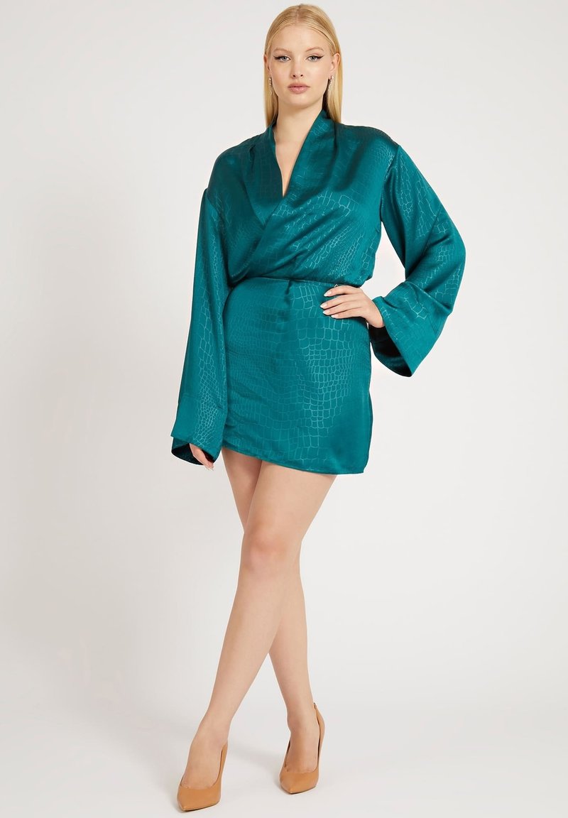 Abito avvolgente in satin color teal con motivo coccodrillo, scollatura a V profonda, maniche larghe e vita segnato da una cintura, abbinato a scarpe con tacco nude.