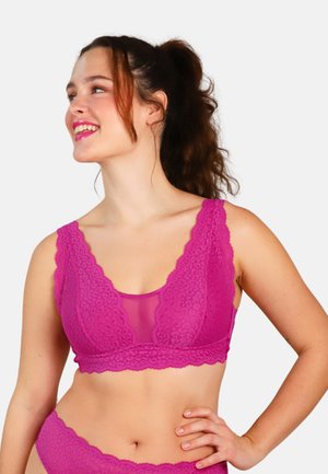 Woman with dark curly hair in a ponytail wearing bright pink lace lingerie, smiling and looking to her left with her hand on her hip.
