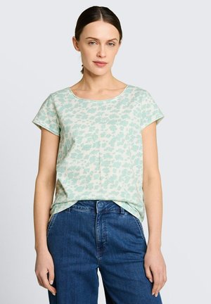 Vrouw met donker haar in een paardenstaart, gekleed in een lichtgroene korte mouw met bloemenpatroon en een blauwe spijkerbroek, staand tegen een effen lichte achtergrond.