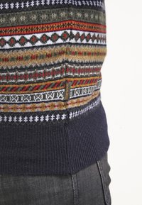 Pull en tricot présentant un motif Fair Isle multicolore dans des tons de vert, rouge, marron et blanc, avec une ceinture côtelée bleu marine foncé.