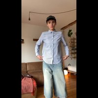Mėlynos ir baltos juostelės mygtukiniai marškiniai, derinami su šviesiai mėlynomis džinsinėmis šortais. Fone matoma sofa ir augalai.