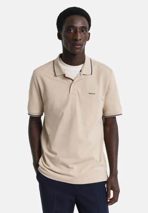 Beige polo skjorte med kontrasterende marineblå krage og mansjetter. Har en liten GANT-logo på venstre bryst i et mykt stoff. Korte ermer.
