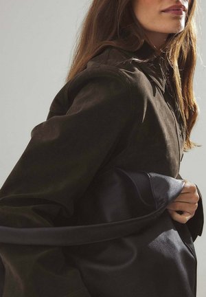 Giacca in suede marrone con collo e chiusura a bottoni, tenuta contro una borsa in pelle nera. Texture liscia, design casual.