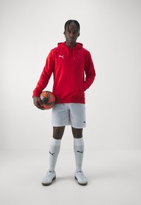 Red hoodie with hood, white shorts, white socks, and sneakers. Holding an orange and black patterned soccer ball. Plain background.