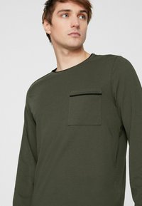 Langärmliges Hemd aus olivgrünem Baumwollstoff, mit einer vorderen Tasche und einem kontrastierenden schwarzen Reißverschluss, rundem Ausschnitt und lockerem Schnitt.