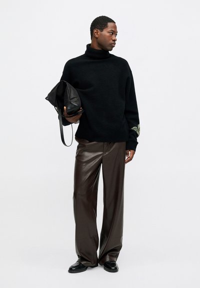 Bărbat îmbrăcat cu pulover negru cu guler înalt, pantaloni largi din piele maro, pantofi negri, ținând o geantă din piele neagră pe un fundal alb.