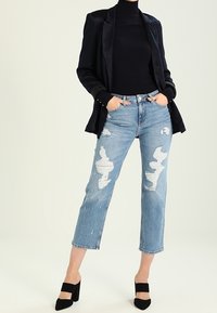Jeans in denim blu con dettagli strappati e una vestibilità dritta, abbinati a un dolcevita nero aderente e a un blazer nero. Scarpe con tacco nere.