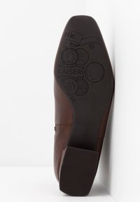 La semelle en cuir marron de la chaussure présente des logos embossés et des motifs complexes, avec un talon plat et un caoutchouc texturé pour une meilleure adhérence.