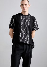 T-shirt preta de mangas curtas com um gráfico de corrente e texto em branco. Corte descontraído com uma textura suave. Usada com calças pretas.