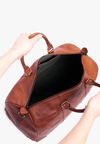 Braune Lederreisetasche mit Reißverschluss oben, zwei Griffen und geräumigem Innenraum aus schwarzem Stoff. Verfügt über Lederakzente und Metallbeschläge.