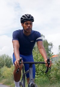 Ciclista con una camiseta y pantalones cortos deportivos de color azul oscuro, con un casco negro, sujetando el manillar de una bicicleta negra, al aire libre con vegetación.