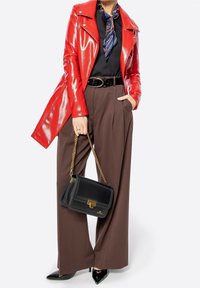 Femme portant une veste en cuir rouge brillante, un chemisier noir, un pantalon marron à jambes larges, des escarpins noirs à bouts pointus, une écharpe bleue à motifs, et tenant un sac à main noir.