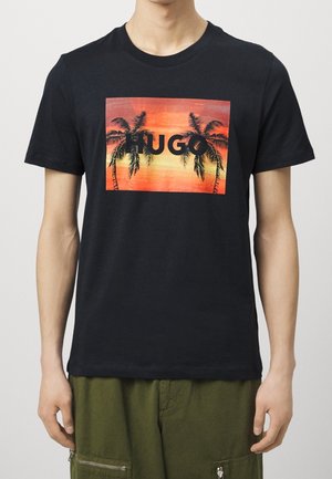 Camiseta de algodón negra con un gráfico colorido de palmeras contra un fondo de atardecer y la palabra "HUGO" en letras negritas negras.