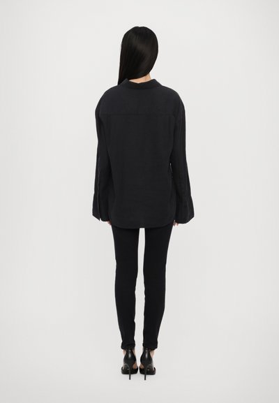 Black linen shirt with long sleeves, relaxed fit, and a rounded hem, paired with black slim-fit pants and heeled shoes.