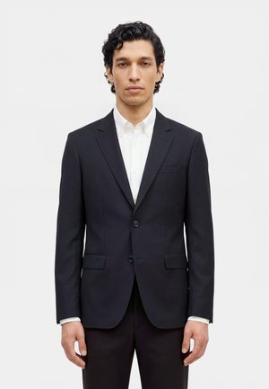 Un homme se tient debout, face à l’objectif, vêtu d’un costume sombre et d’une chemise blanche, sur un fond clair et uni.
