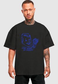 Sort oversized t-shirt lavet af bomuld med et blåt grafisk motiv af en smilende person, der holder pengesedler, med teksten "TJEN PENGE UDEN VENNER."