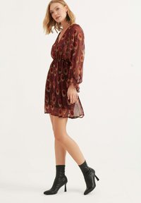 Kurzärmliges Kleid mit tiefem V-Ausschnitt und floralem Muster in Rot und Gelb. Leichtes, transparentes Material. Kombiniert mit schwarzen, hohen Stiefeletten.