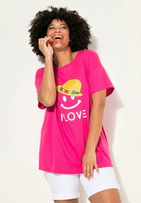 Camiseta de algodón rosa con mangas cortas, que presenta un gráfico de una cara sonriente con un sombrero amarillo y la palabra "AMOR" en blanco.