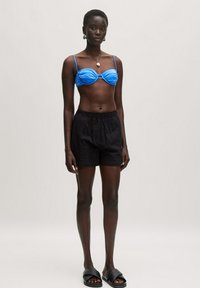 Blaue Bandeau-Bikini-Oberteil mit Raffung, kombiniert mit schwarzen strukturierten Shorts und schwarzen Slide-Sandalen. Schlichtes Design und leichtes Material.